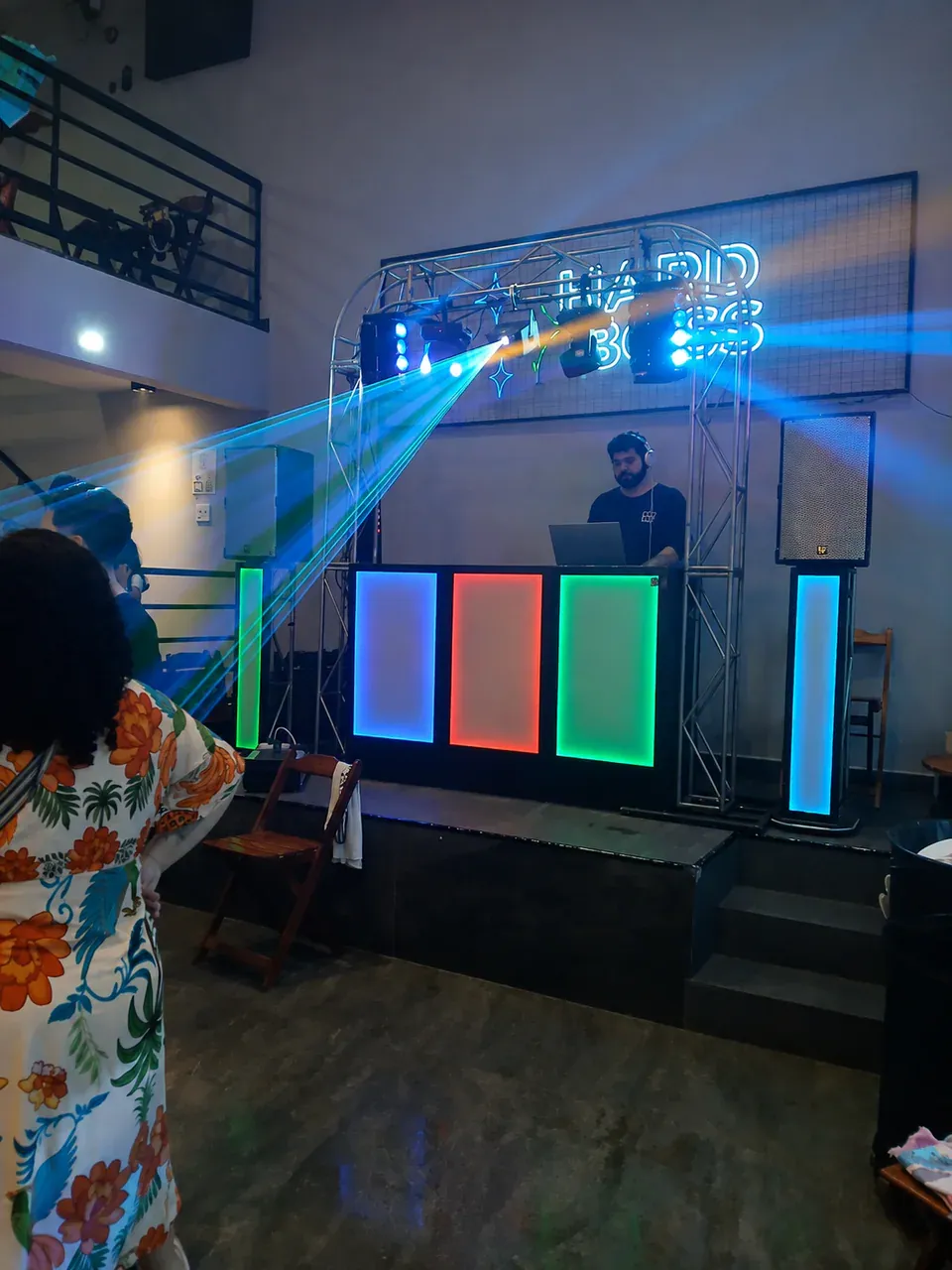 DJ atrás de cabine iluminada com caixas acústicas e feixes de luz.