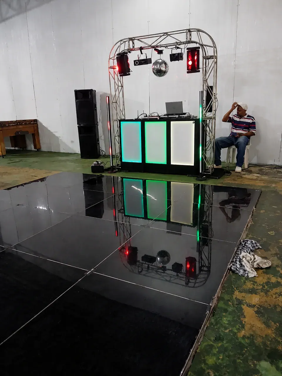 Pista preta refletiva diante do DJ. Piso modular escuro refletindo a cabine iluminada e criando continuidade visual entre música e pista.