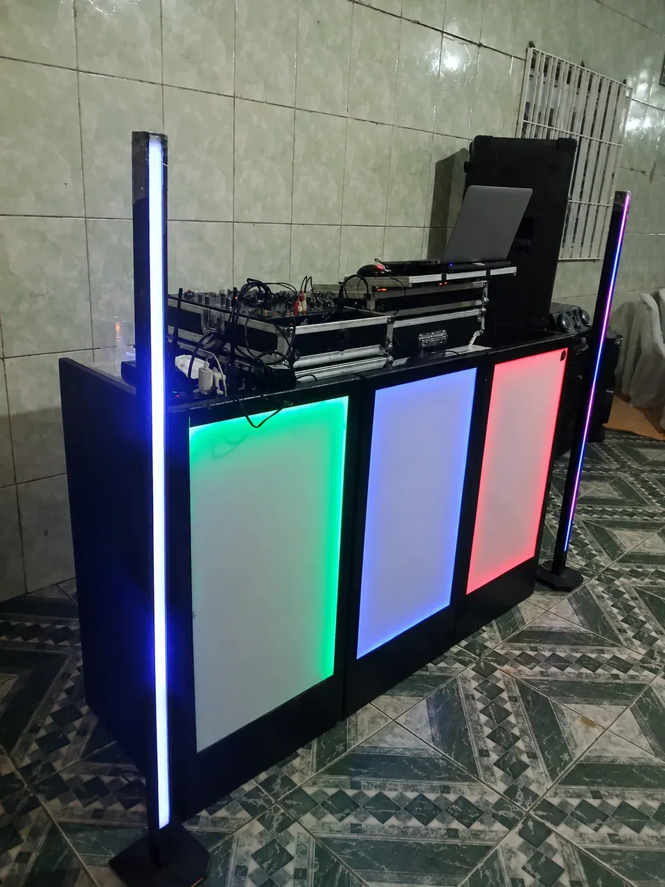 Cabine iluminada com equipamento de DJ. Detalhe da área de operação com controladora, mesa de áudio, notebook e barras luminosas.